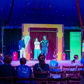 Zwei Personen stehen auf einer Bühne im Zirkus, während das Publikum in blauen T-Shirts zusieht.