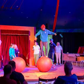 Zirkusaufführung mit einem Artisten, der auf zwei großen roten Bällen balanciert, umgeben von Zuschauern und anderen Darstellern.