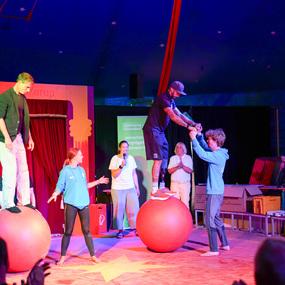 Aufführung im Zirkus: Künstler stehen auf großen Bällen und interagieren mit dem Publikum in festlichem Zelt.