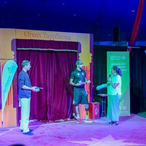 Zwei Personen auf einer Bühne im Zirkuszelt, eine spricht ins Mikrofon, während der andere aufmerksam zuhört.