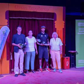 Vier Personen stehen auf einer Zirkusbühne, präsentieren Auszeichnungen vor einem roten Vorhang und Bannern von Mettmann-Sport.