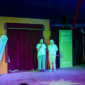 Zwei Personen stehen auf einer Zirkusbühne, umgeben von bunten Vorhängen und Werbebannern in einem Zelt.