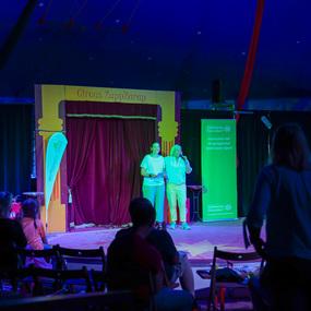 Zwei Personen stehen auf einer Bühne im Zirkuszelt und präsentieren vor Publikum, umgeben von Dekoration und Banner.