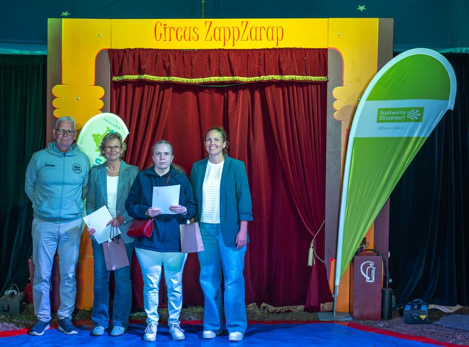 Vier Personen stehen vor einem roten Vorhang im Zirkuszelt von "Circus ZappZarap", halten Dokumente in der Hand.