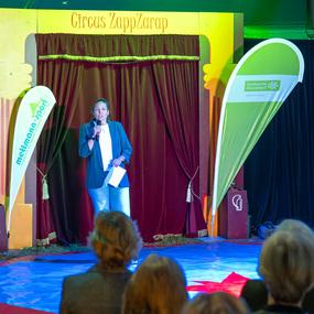Eine Frau spricht am Mikrofon vor einem roten Vorhang im Zelt des Zirkus ZappZarap, Publikum sitzt im Vordergrund.