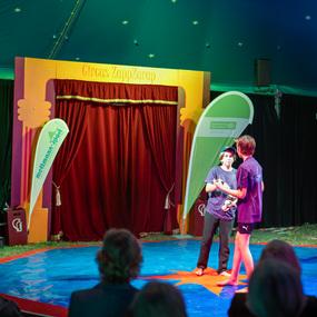 Junge Artisten in einem Zirkuszelt üben Jonglieren auf der blauen Bühne mit rotem Vorhang im Hintergrund.