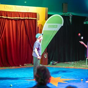Zwei Kinder im Zirkus werfen jonglierend Bälle auf einer bunten Matte vor einem roten Vorhang.