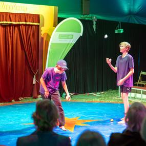 Zwei Kinder in T-Shirts jonglieren auf einer blauen Zirkusfläche, während Zuschauer im Hintergrund zuschauen.