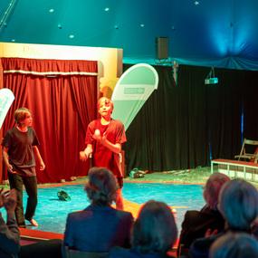 Zwei Jongleure führen in einem Zirkuszelt mit Publikumsbeteiligung eine Darbietung auf.