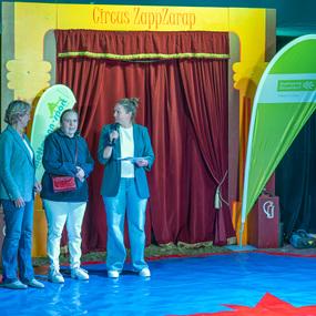 Drei Frauen stehen auf einer bunten Zeltbühne. Eine spricht ins Mikrofon, während die anderen zuhören.