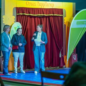 Drei Frauen stehen auf einer Zirkusbühne, während eine von ihnen ein Mikrofon hält und spricht.