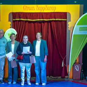 Vier Personen stehen vor einem roten Vorhang im Zirkuszelt von "Circus ZappZarap", halten Dokumente in der Hand.