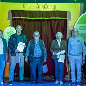 Fünf Personen stehen lächelnd vor einem roten Vorhang im Circus ZappZarap, halten Urkunden und kleine Geschenktüten.