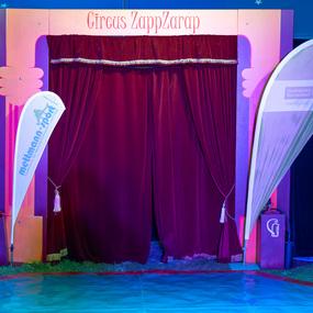 Eingang des Circustheaters „Circus ZappZarap“ mit roten Vorhängen und Werbebannern in buntem Licht.