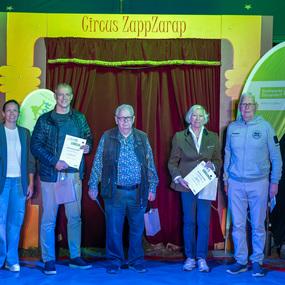 Fünf Personen stehen vor einem roten Vorhang im Zirkuszelt und halten Urkunden in den Händen.