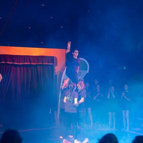 Akrobatin steht auf den Händen mehrerer Mitspieler in einem Zirkus, umgeben von buntem Nebel und Publikum.