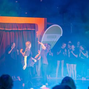 Aufführung im Zirkus, mit jungen Tänzern auf einer Bühne und Zuschauern im Nebel. Farbige Lichter und ein Vorhang im Hintergrund.