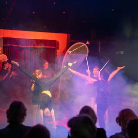 Aufführung im Zelt mit mehreren Künstlerinnen, die Akrobatik und einen Stabantanz in buntem Nebel zeigen.