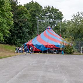 Bunte Zirkuskuppel in einem Park, umgeben von Bäumen und einer kleinen Menschenmenge auf einem Asphaltplatz.