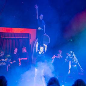 Zwei Mädchen führen eine Akrobatik-Nummer auf, während andere Tänzerinnen im Nebel zuschauen. Die Bühne ist beleuchtet.