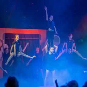 Acht Akrobatinnen führen eine Menschenpyramide in einem Zirkuszelt auf, umgeben von buntem Rauch.