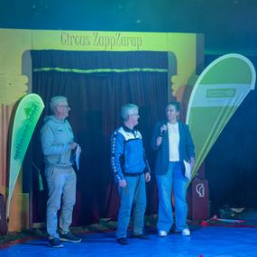 Drei Personen stehen auf einer blauen Fläche vor einem Circus-Zelt und sprechen ins Mikrofon. Hinter ihnen ist eine Vorhang.