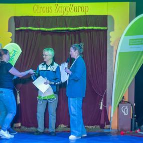 Vier Personen stehen auf einer Zirkusbühne, Übergabe eines Dokuments vor einem bunten Zirkushintergrund.