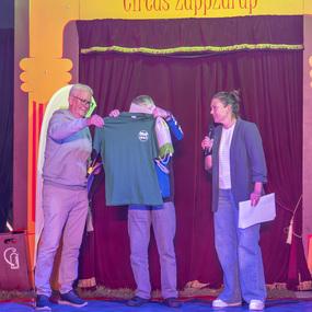 Zwei Männer halten ein grünes T-Shirt in der Mitte einer Zirkusbühne, während eine Frau mit Mikrofon spricht.