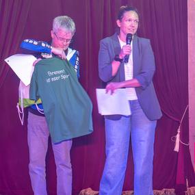 Ein Mann hält ein grünes T-Shirt mit dem Text „Ehrenamt ist unser Sport“ während eine Frau mit Mikrofon spricht.