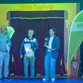 Drei Personen stehen auf einer Bühne im Zelt des Circus ZappZarap, während eine Frau ins Mikrofon spricht.