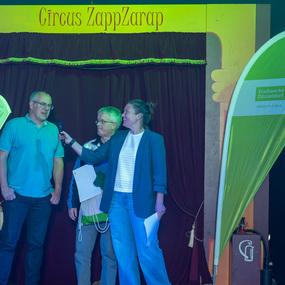 Zwei Männer und eine Frau stehen auf der Bühne des Circus ZappZarap, während sie ein Mikrofon benutzen.