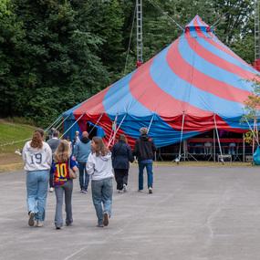 Gruppe von Menschen geht auf einen bunten Zirkuswagen zu, umgeben von Bäumen auf einem gepflasterten Weg.