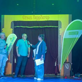 Drei Personen stehen auf einer B&uuml;hne im Zirkus ZappZarap, w&auml;hrend eine Frau mit Mikrofon interviewt.
