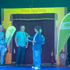 Drei Personen stehen auf einer Zirkusb&uuml;hne, w&auml;hrend eine Frau Mikrofon und Notizen in der Hand h&auml;lt.