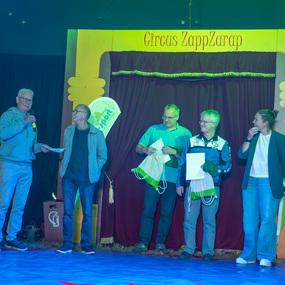 Vier Personen stehen auf einer B&uuml;hne des Cirque ZappZarap und h&ouml;ren einem Redner zu, w&auml;hrend sie Taschen und Papiere halten.