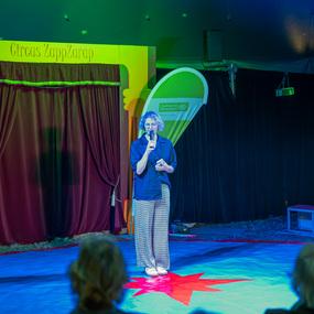 Eine Frau steht auf einer bunten Zirkusfl&auml;che und spricht mit Mikrofon, im Hintergrund der Zirkusvorhang und Zuschauer.