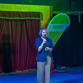 Frau mit Mikrofon steht auf einer blauen Fl&auml;che vor einem Theatervorhang in einem Zirkuszelt mit bunten Werbetransparenten.