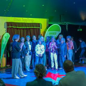 Eine Gruppe von Frauen steht im Zirkus, w&auml;hrend eine Moderatorin spricht. Bunte Lichter erleuchten die B&uuml;hne.