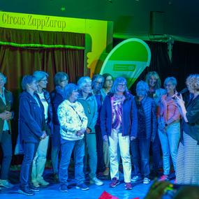 Gruppe von Frauen steht in einem Zelt, w&auml;hrend eine Teilnehmerin mit Mikrofon spricht, im Hintergrund die Kulisse des Circus ZappZarap.