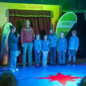 Kinder stehen auf einer Zirkusb&uuml;hne, w&auml;hrend ein Erwachsener sie anspricht. Zirkusbanner im Hintergrund sichtbar.