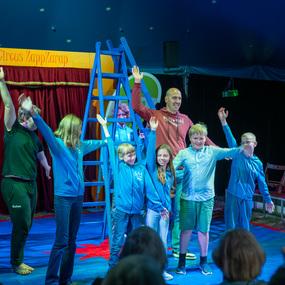 Gruppe von Kindern und einem Erwachsenen, die auf einer Zirkusbühne den Zuschauern zuwinken.