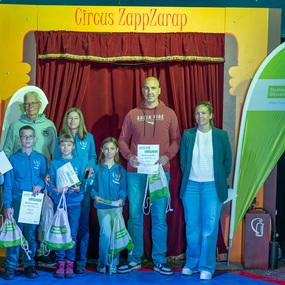 Gruppe von sechs Kindern und Erwachsenen bei einer Preisverleihung im Zirkus ZappZarap, allesamt l&auml;cheln und halten Urkunden.