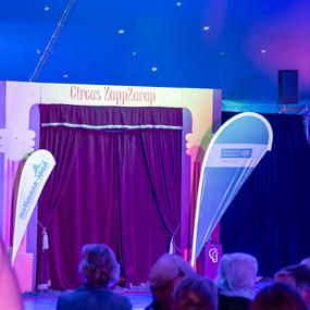 Bühne im Zelt des Circus ZappZarap mit rotem Vorhang und zwei Werbebannern, umgeben von einem Publikum.