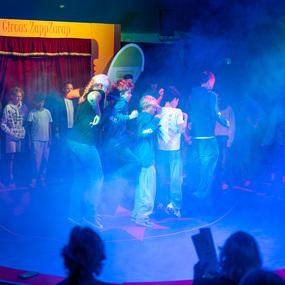 Gruppenszene im Zirkus mit Kindern, die auf dem Boden stehen, umgeben von buntem Rauch und einer B&uuml;hne im Hintergrund.