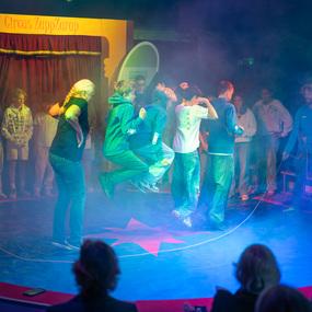 Gruppe von Kindern springt auf einer bunten B&uuml;hne im Zirkus, umgeben von Rauch und Zuschauer.