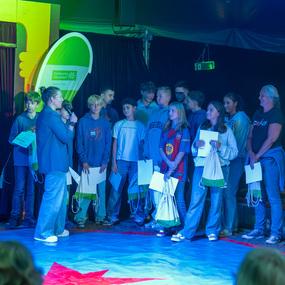 Eine Gruppe von Jugendlichen steht auf einer B&uuml;hne, w&auml;hrend ein Moderator spricht und die Anwesenden beobachtet.