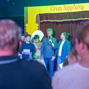 Auf der B&uuml;hne des Circus ZappZarap stehen f&uuml;nf Personen; eine Frau interviewt einen Mann, w&auml;hrend das Publikum zuschaut.