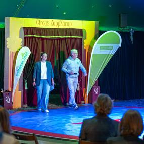 Zwei Moderatoren treten durch den Eingang eines Zeltes mit dem Schriftzug „Circus ZappZarap“ auf eine bunte Bühne.