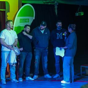 Vier M&auml;nner stehen zusammen auf einer B&uuml;hne, w&auml;hrend eine Frau mit Mikrofon sie anspricht. Im Hintergrund ist ein Banner.