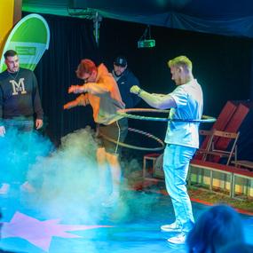 Vier M&auml;nner stehen in einer bunten Zirkusumgebung, w&auml;hrend einer mit einem Hula-Hoop-Reifen jongliert.